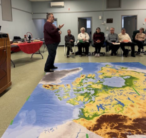 A man gestures while speaking to a seated group gathered around a large, colourful floor map of Canada.