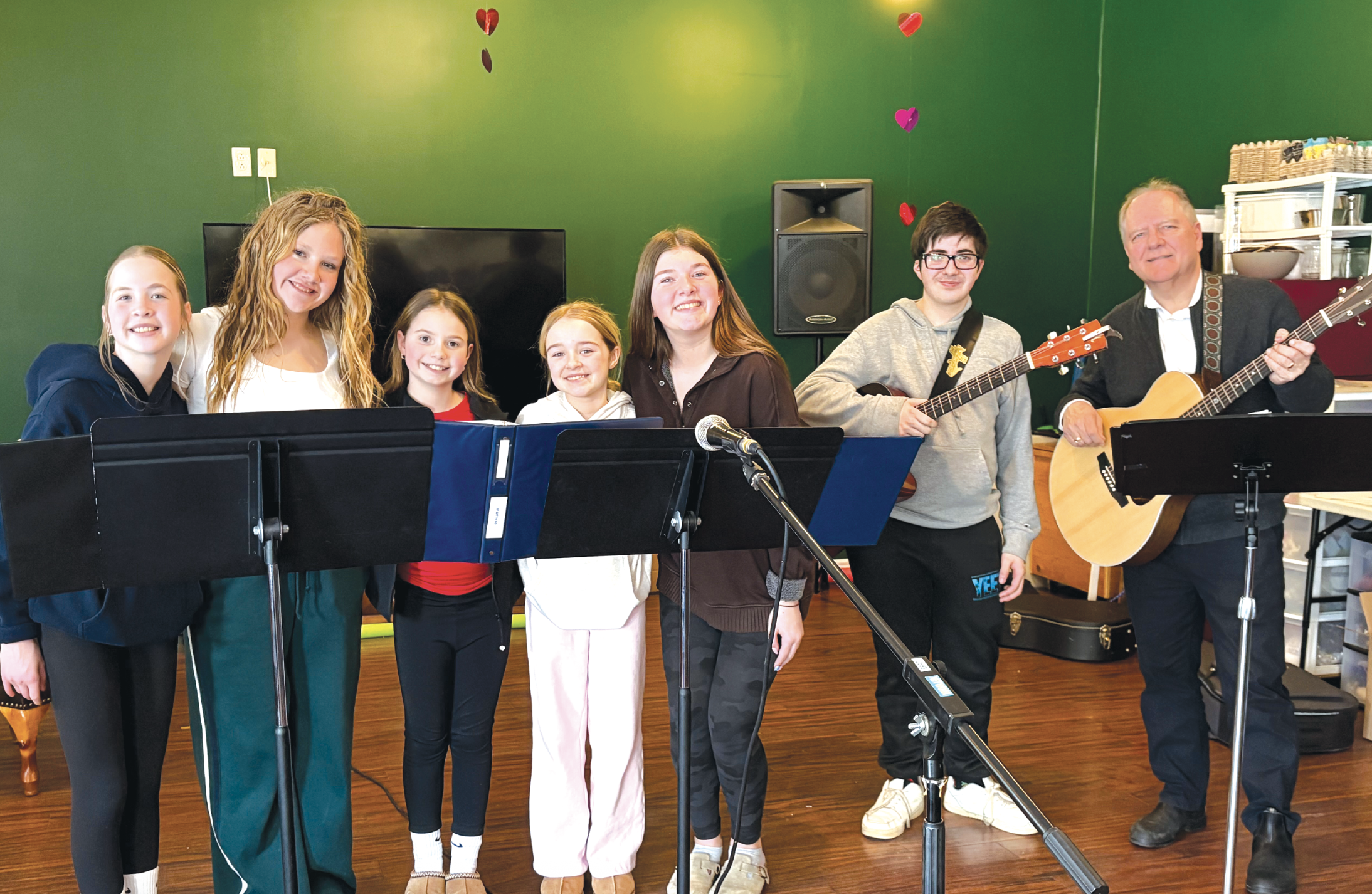 a group of people, young and old, stand together with microphones and a guitar in a green room