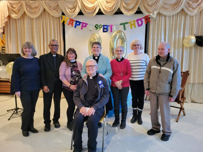 Group photo of eight people celebrating a 90th birthday in a decorated hall.