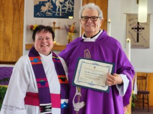 Two priests at the front of a church: on the right a man dressed in a purple chasuable holding a certificate; on the left a woman dressed in a white alb and a colourful stole