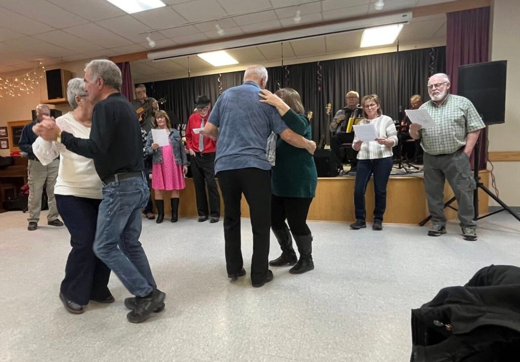 Several couples dancing in a community centre while a live band performs on a stage in the background.