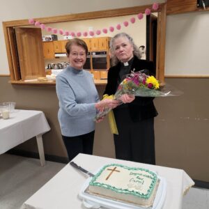 two women, one dressed in black as a priest, hold a cake