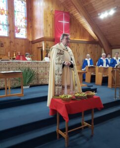A priest in ornate cream-colored vestments stands before an altar with baskets of palm fronds on a red-clothed table.