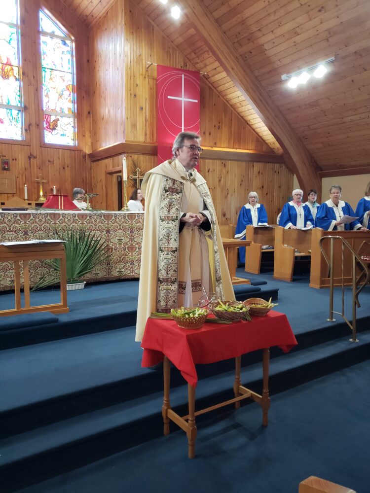 A priest in ornate cream-colored vestments stands before an altar with baskets of palm fronds on a red-clothed table.