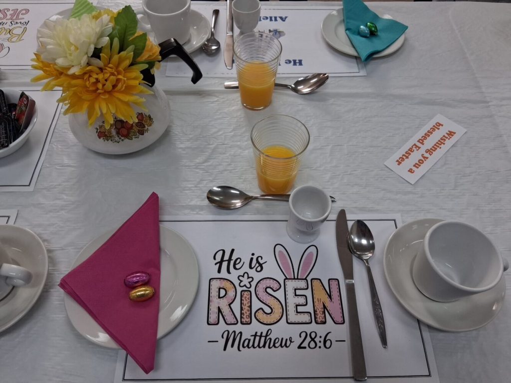 A close-up of an Easter breakfast table setting featuring a "He is Risen" placemat, coffee cups, orange juice, and small chocolate eggs.
