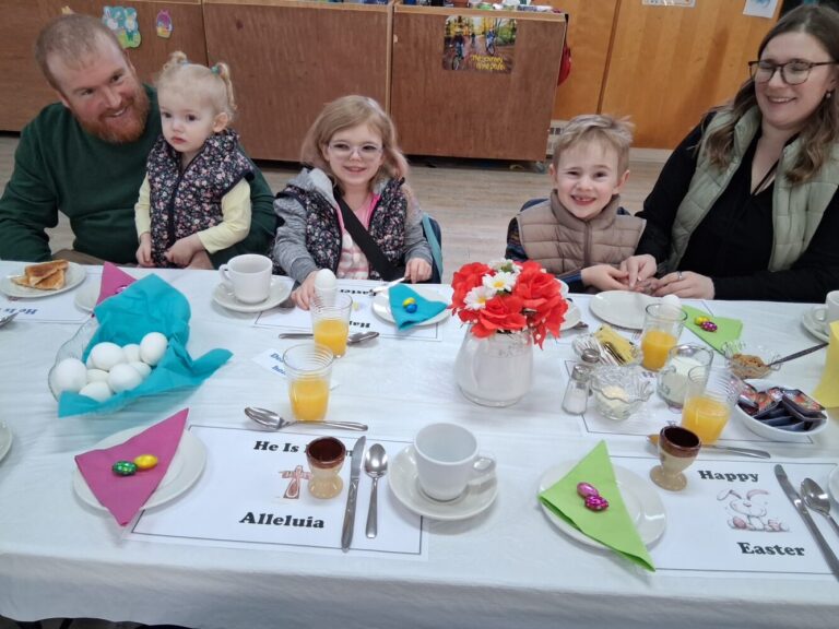 A family with three young children sits at a breakfast table decorated with Easter placemats, orange juice, and colorful napkins.