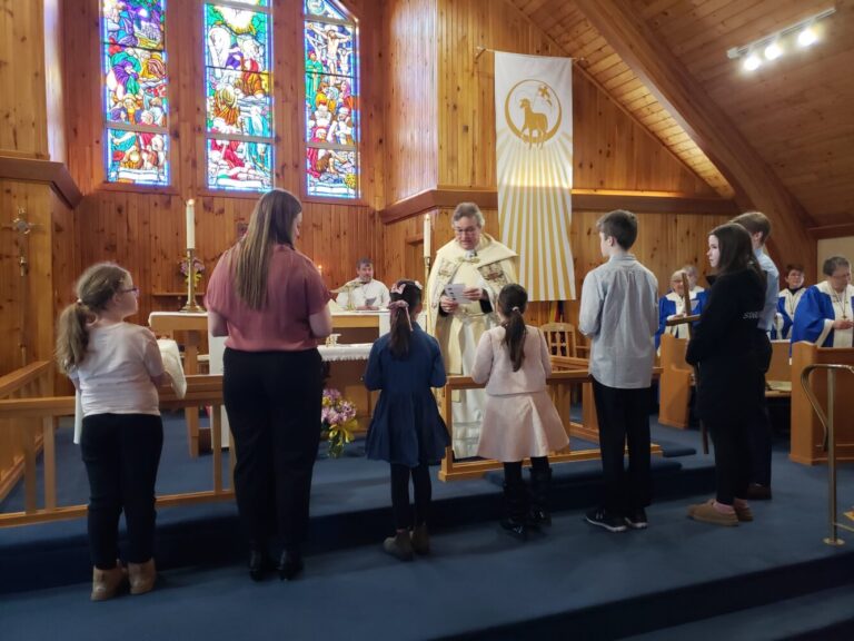 A group of children and adults stand at the front of a church while a priest speaks during a service.