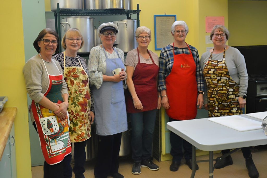 Six women wearing festive aprons stand together in a kitchen.