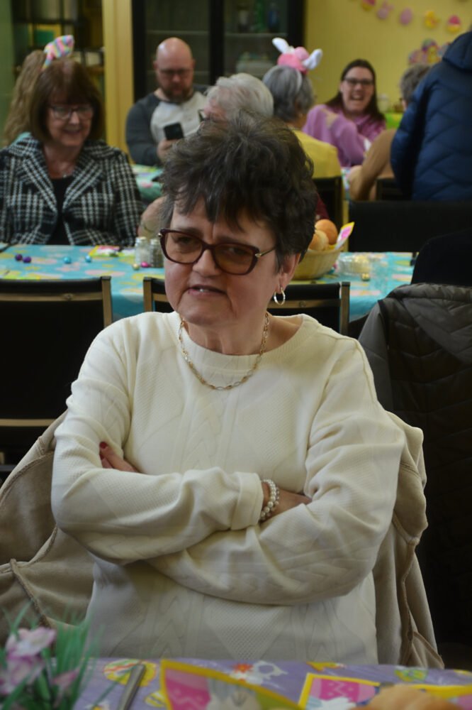 A woman in a white sweater and glasses sitting at a table in a hall decorated for Easter