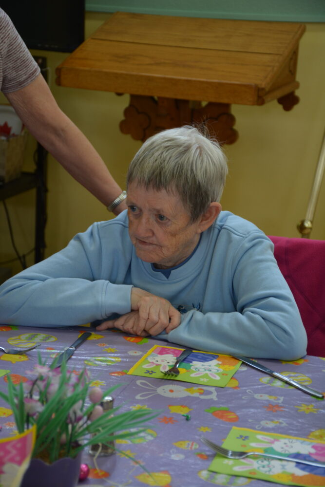 An elderly woman in a blue sweatshirt sits at a table with Easter decorations