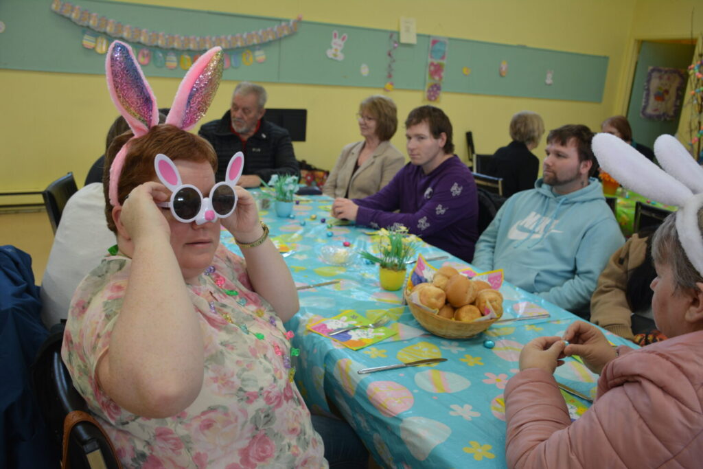 A group of people sit at a dining table; one woman in the foreground wears bunny ears and bunny-shaped sunglasses.