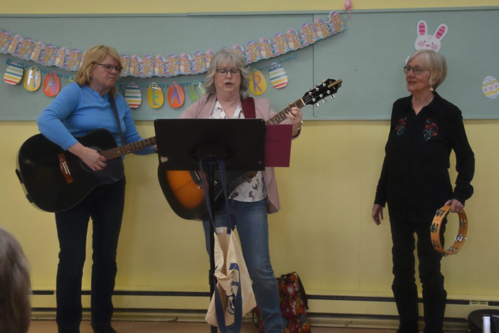 Two women play acoustic guitars while a third woman holds a tambourine in front of an Easter banner.