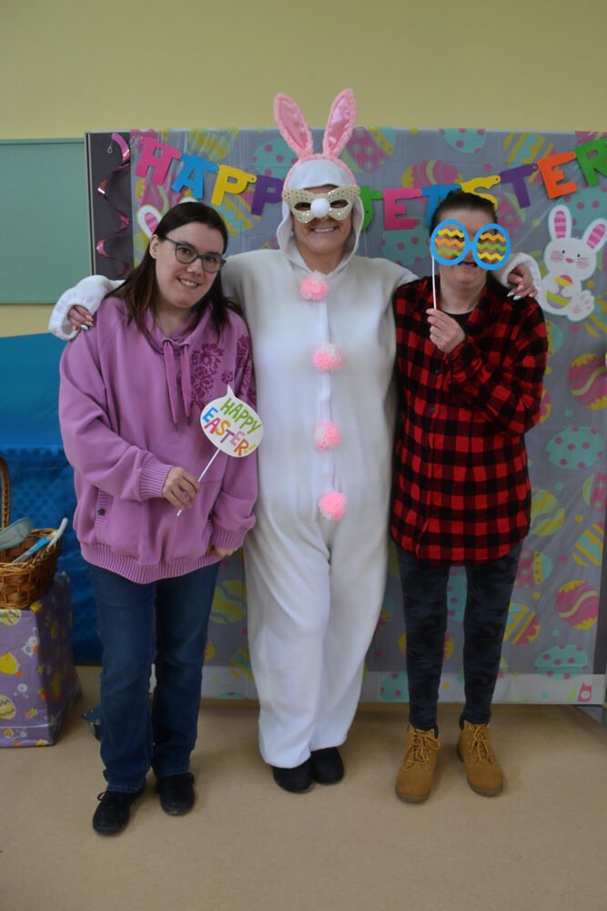 Two women pose for a photo with a person in a white bunny costume in front of a "Happy Easter" backdrop.