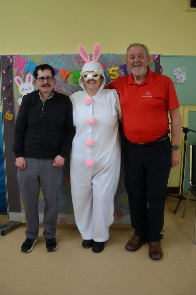 Two men pose with a person in a white bunny costume in front of a festive backdrop.