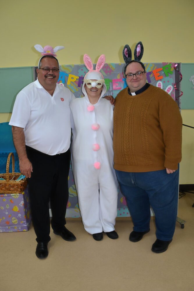 Two men pose with a person in a white bunny costume in front of a festive backdrop.