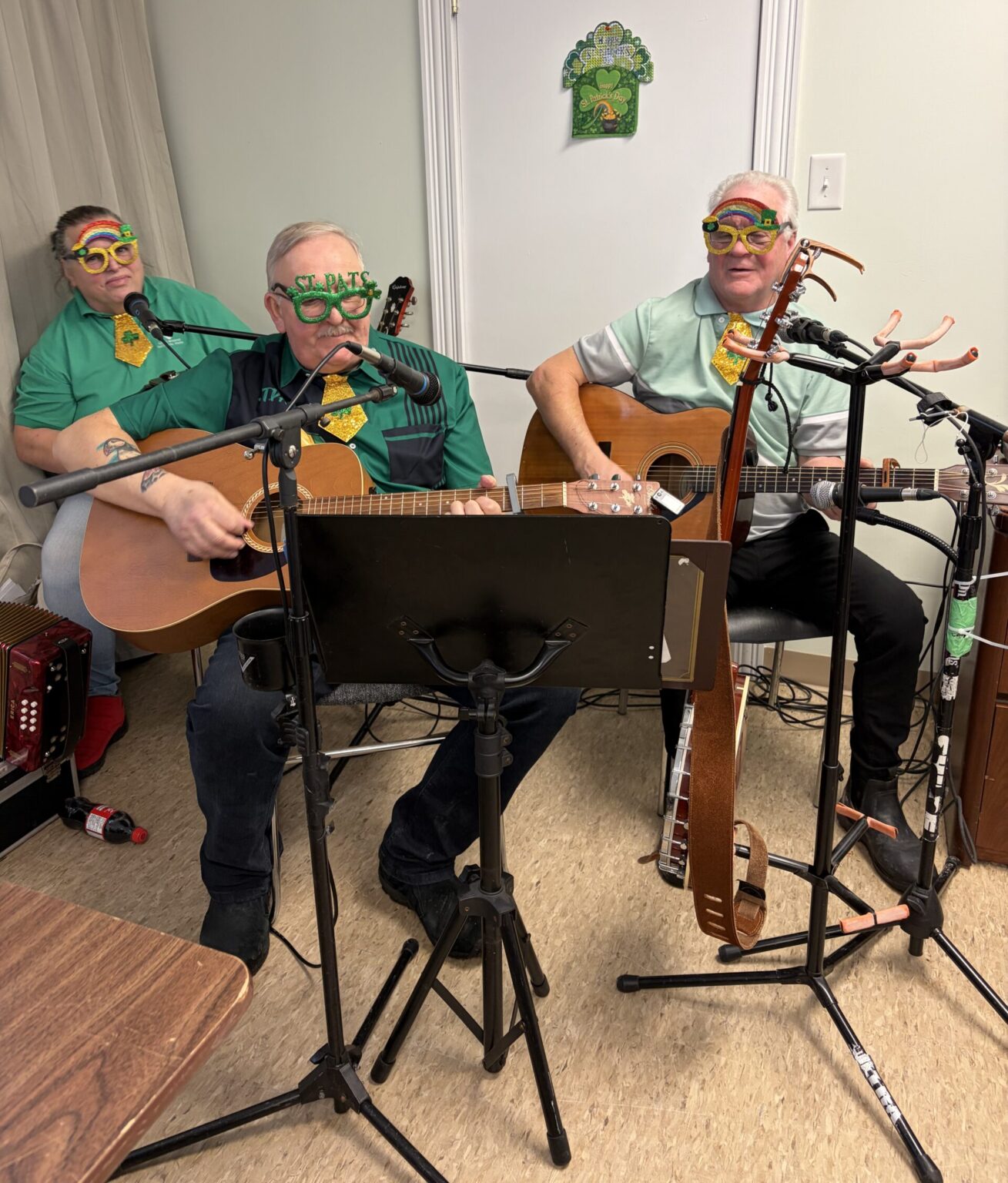 Three musicians wearing green shirts and festive St. Patrick's Day glasses perform with guitars and a microphone.
