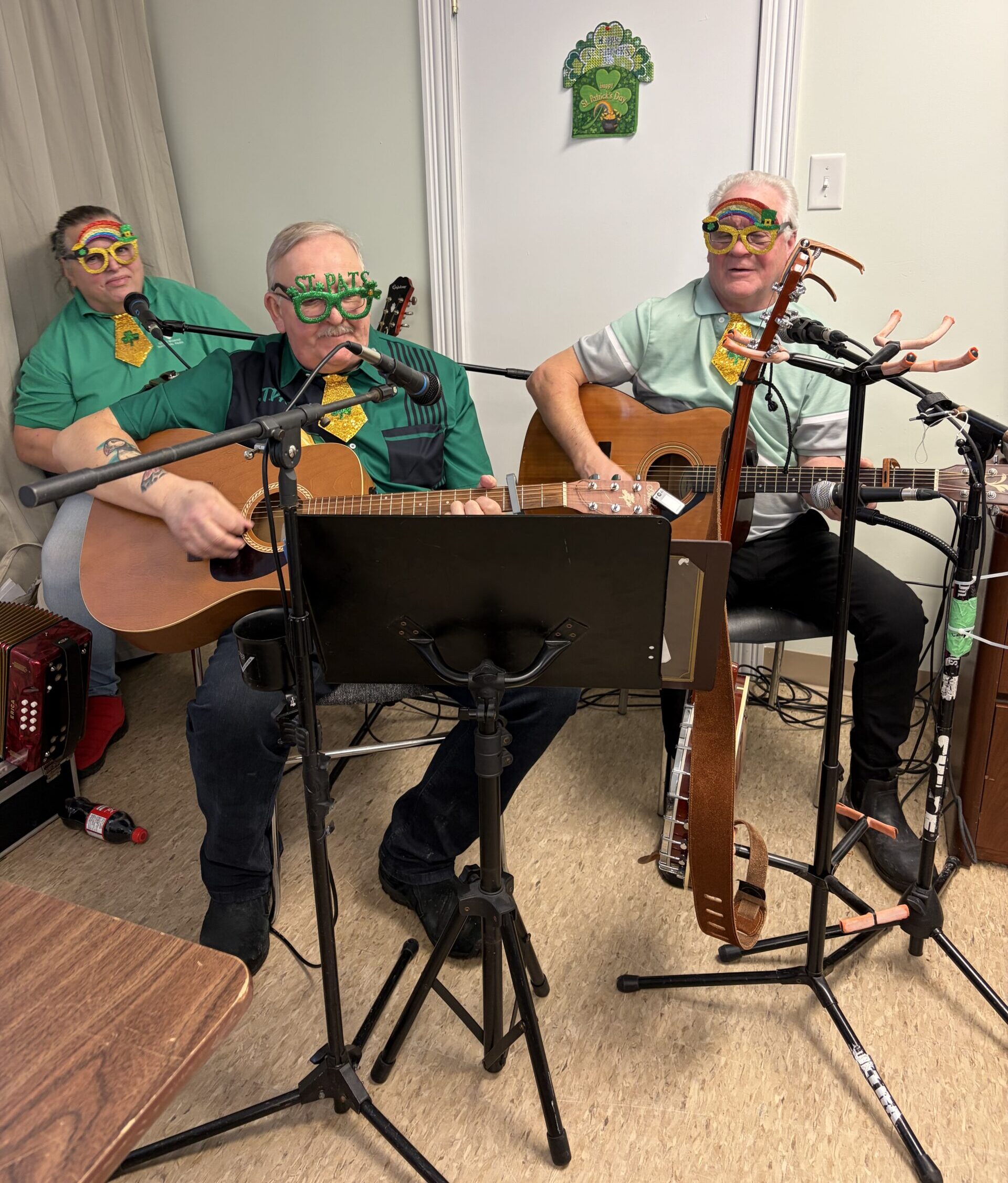 Three musicians wearing green shirts and festive St. Patrick's Day glasses perform with guitars and a microphone.