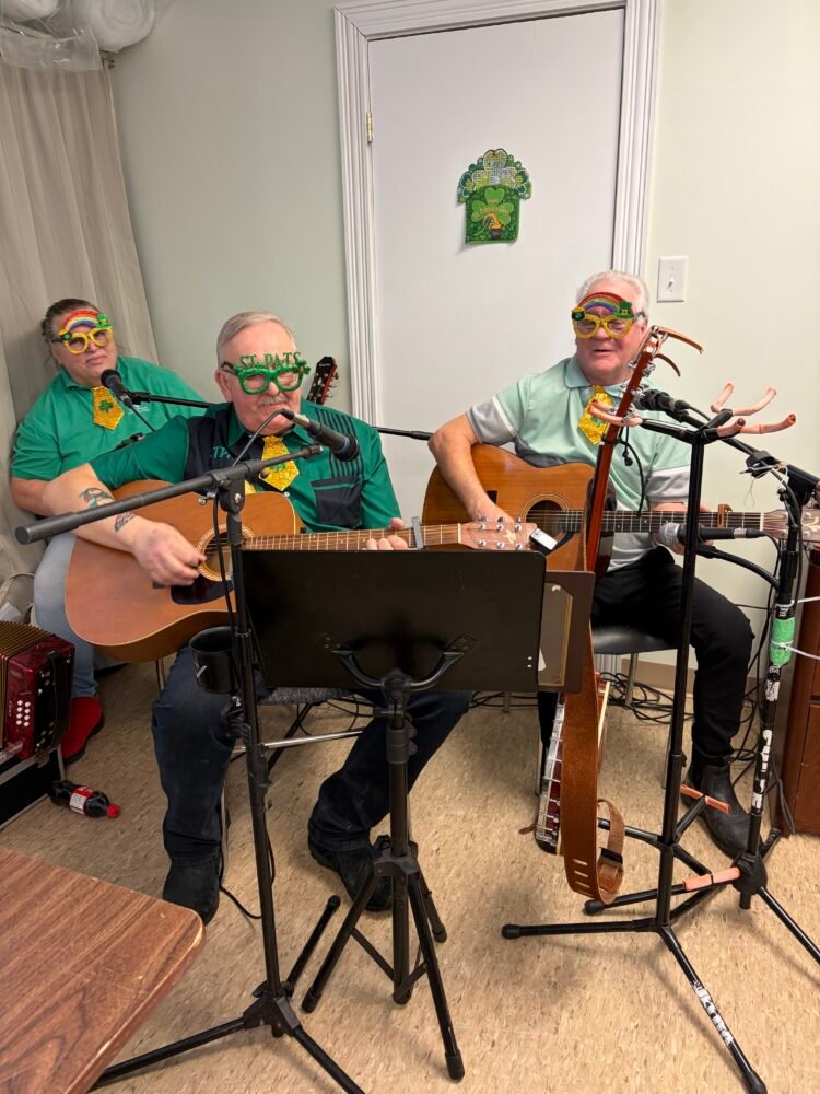 Three musicians wearing green shirts and festive St. Patrick's Day glasses perform with guitars and a microphone.