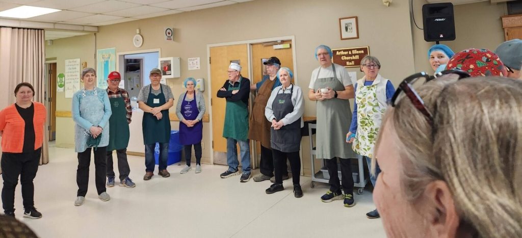 A line of volunteers in aprons and hairnets stand ready in a hall labeled "Arthur & Eileen Churchill Hall."