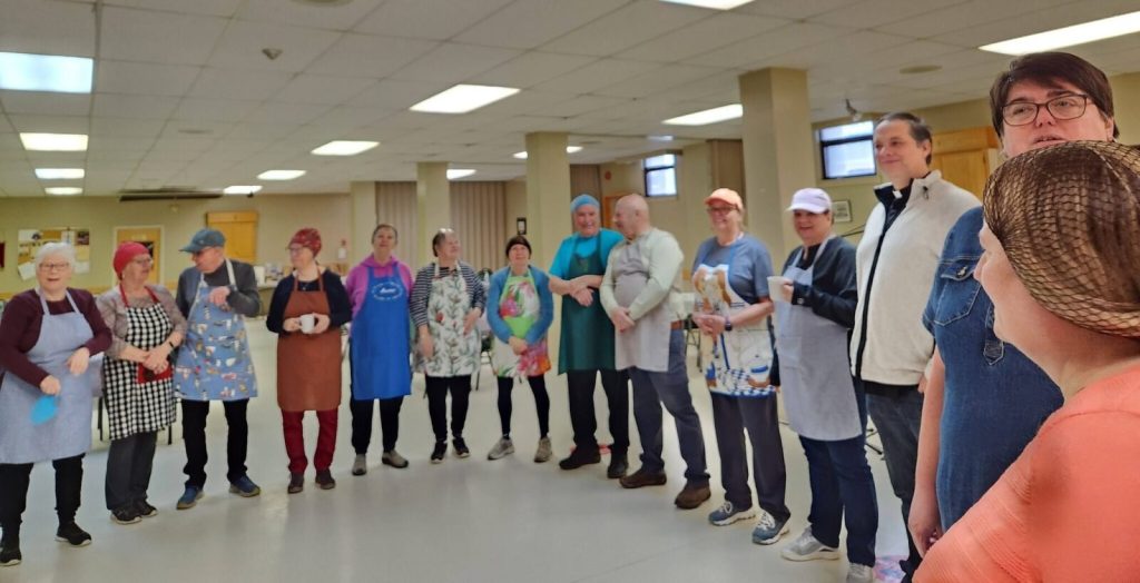 A large group of volunteers wearing colorful aprons and hairnets stand in a circle in a community hall.