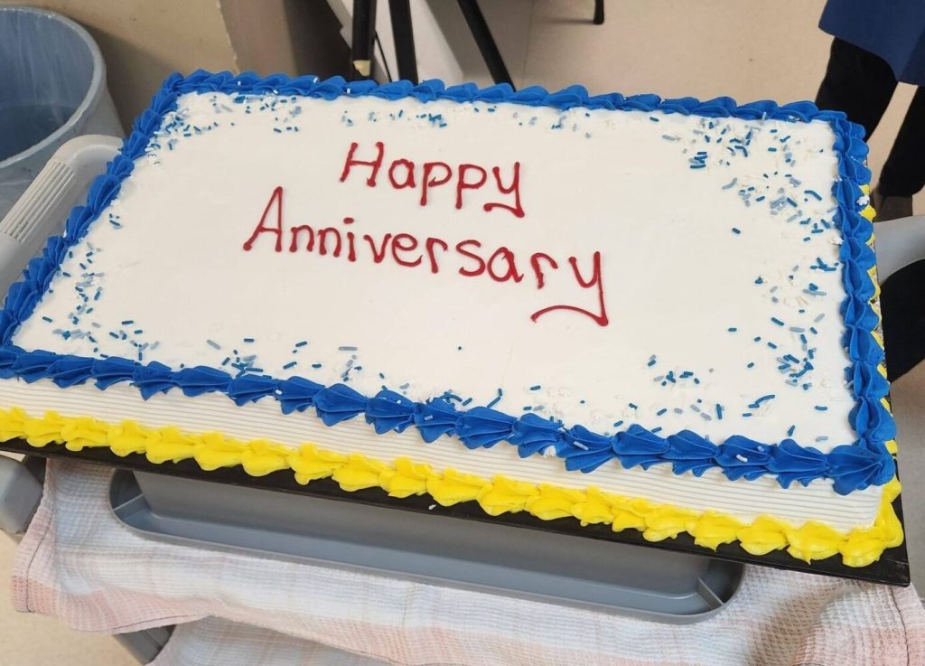 A large rectangular sheet cake with white frosting, blue and yellow borders, and "Happy Anniversary" written in red icing.