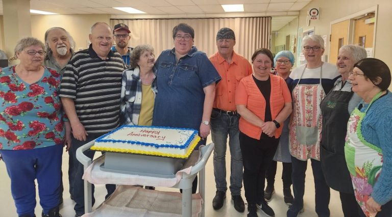 A group of people posing together behind a "Happy Anniversary" cake on a rolling cart.