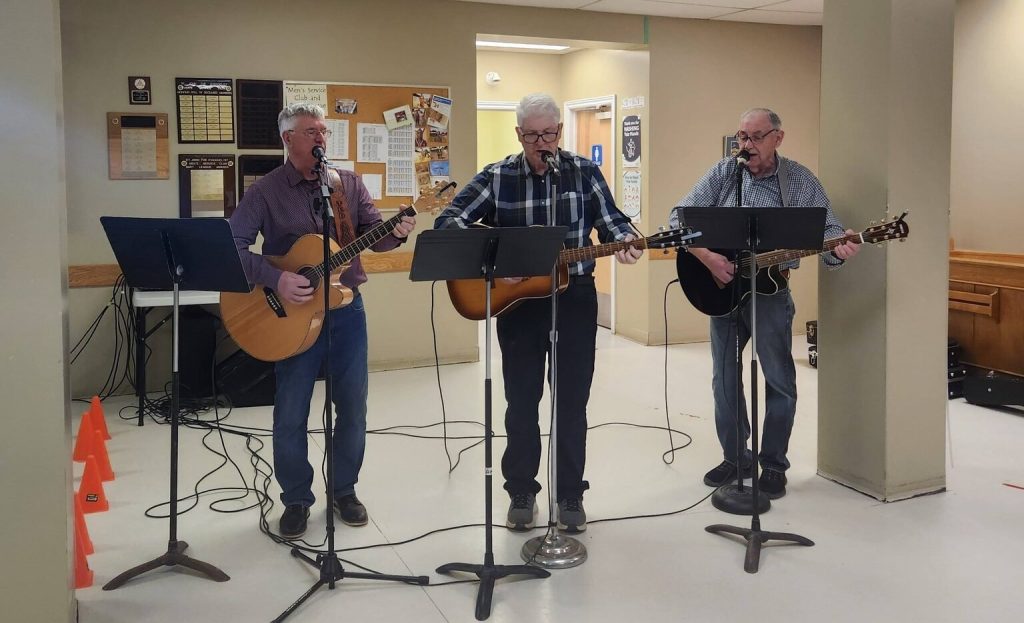 Three men with acoustic guitars and microphones perform music in front of a community bulletin board.