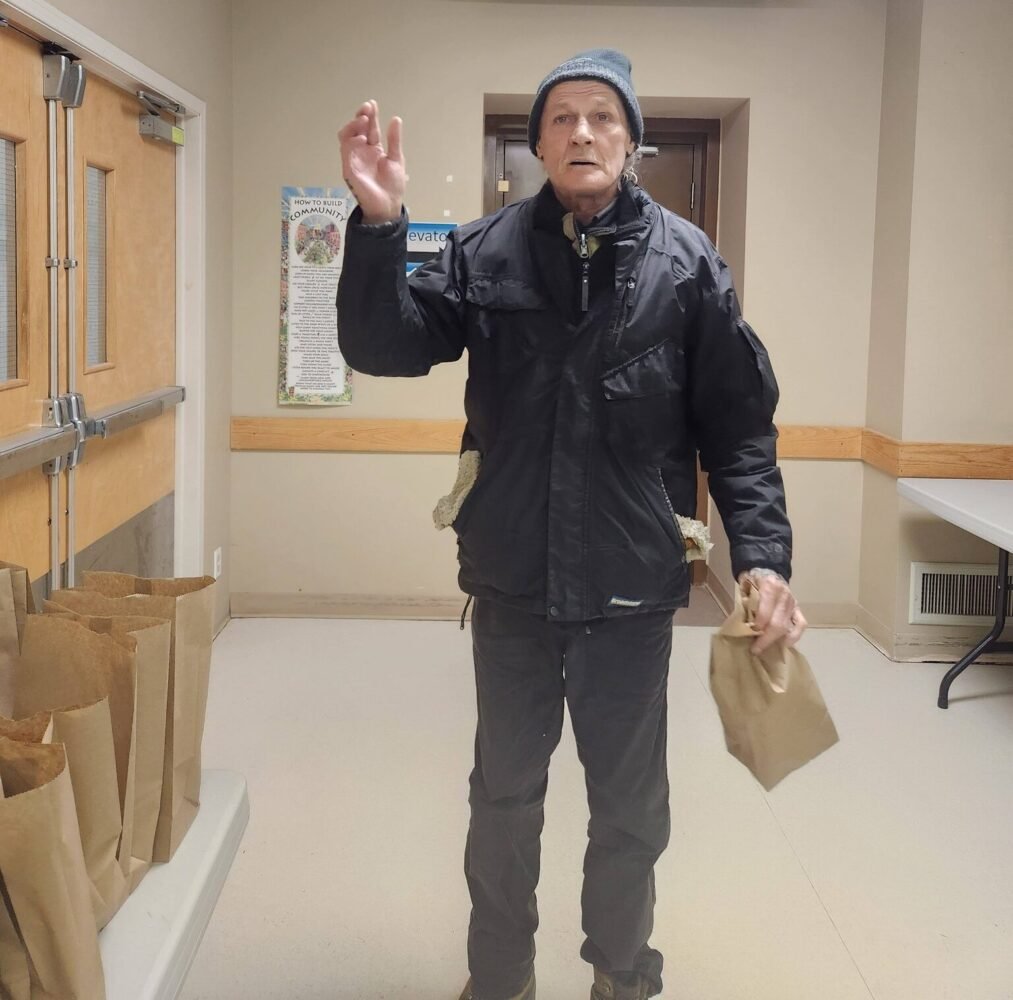 A man in a black jacket and beanie waves while holding a brown paper bag; more bags line a table nearby.