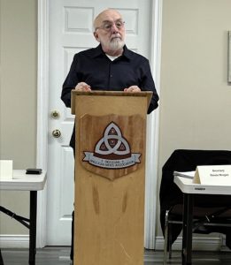 A man with a white beard stands behind a wooden lectern featuring a shield that reads "Diocesan Anglican Men's Association."