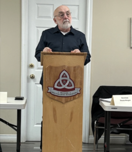A man with a white beard stands behind a wooden lectern featuring a shield that reads "Diocesan Anglican Men's Association."