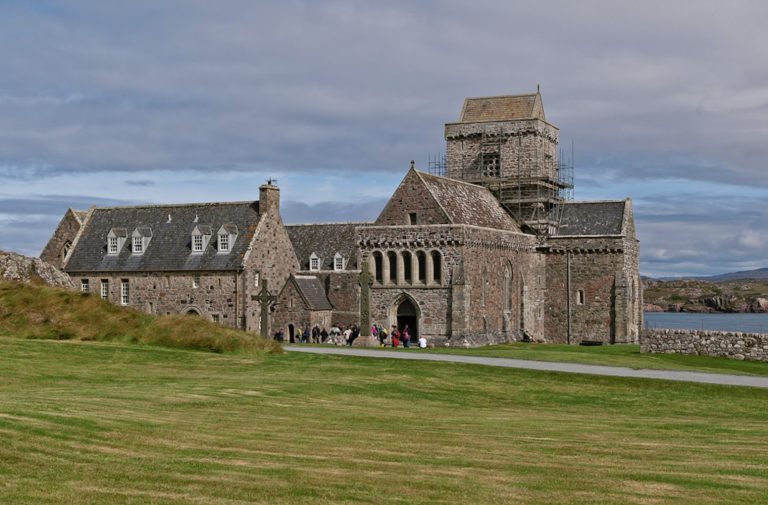 A historic stone abbey with a square tower under scaffolding stands on a grassy field near the coast.