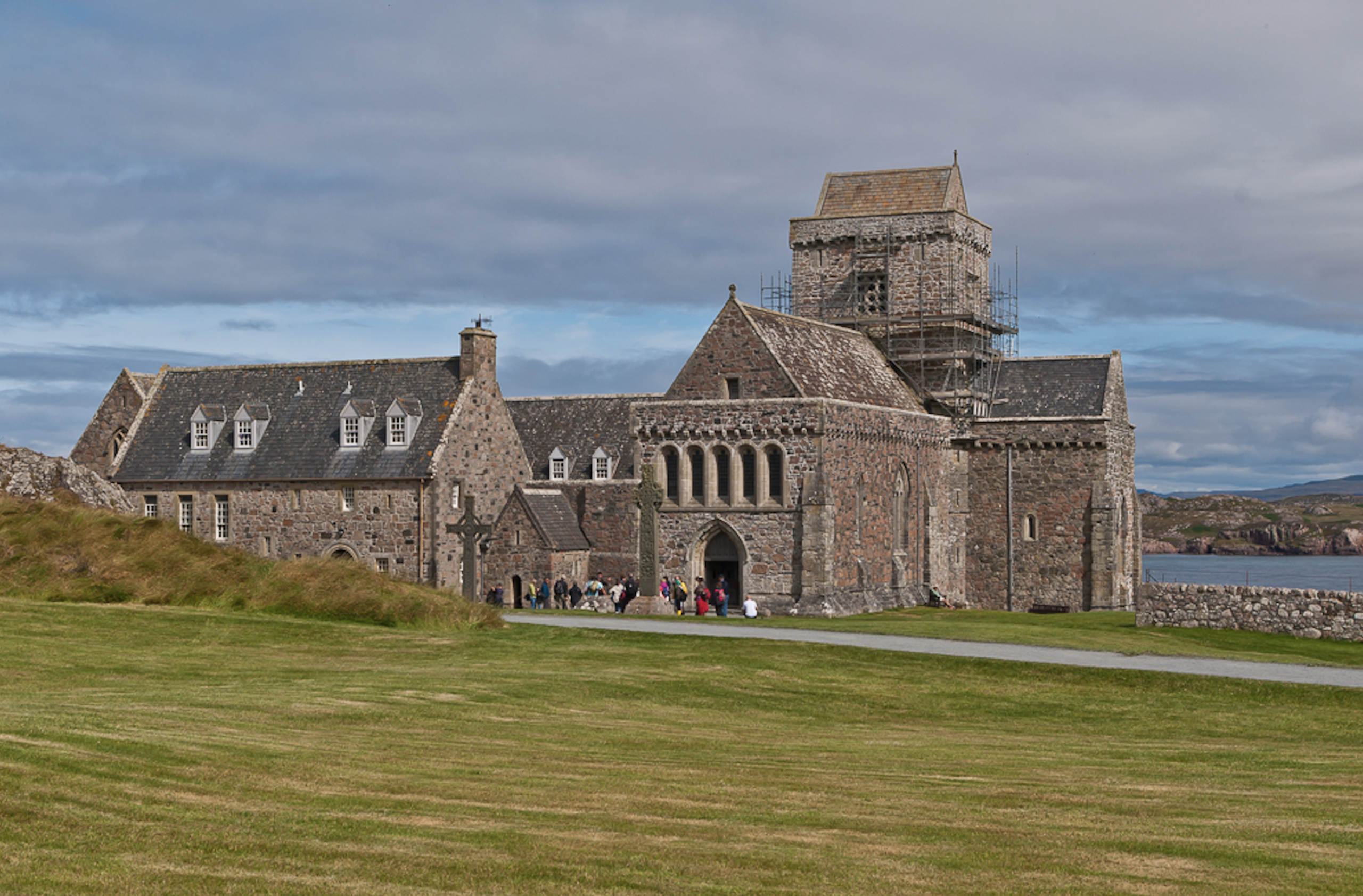 A historic stone abbey with a square tower under scaffolding stands on a grassy field near the coast.