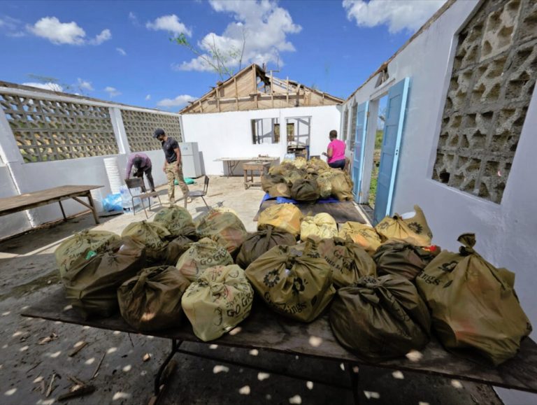 Dozens of filled yellow and brown bags sit on tables inside a roofless, damaged building being used for aid distribution.