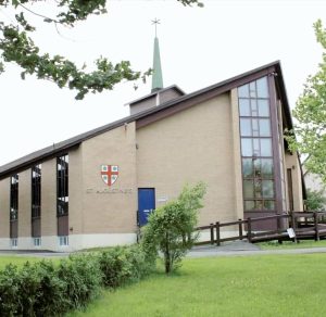Modern tan brick church with a green steeple, tall stained-glass windows, and the name "St. Augustine's" below a red and white crest.