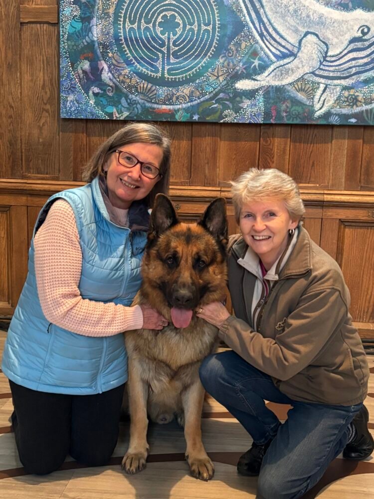 Two women smiling while kneeling with a large German Shepherd in front of wood paneling and a blue nautical-themed mural.