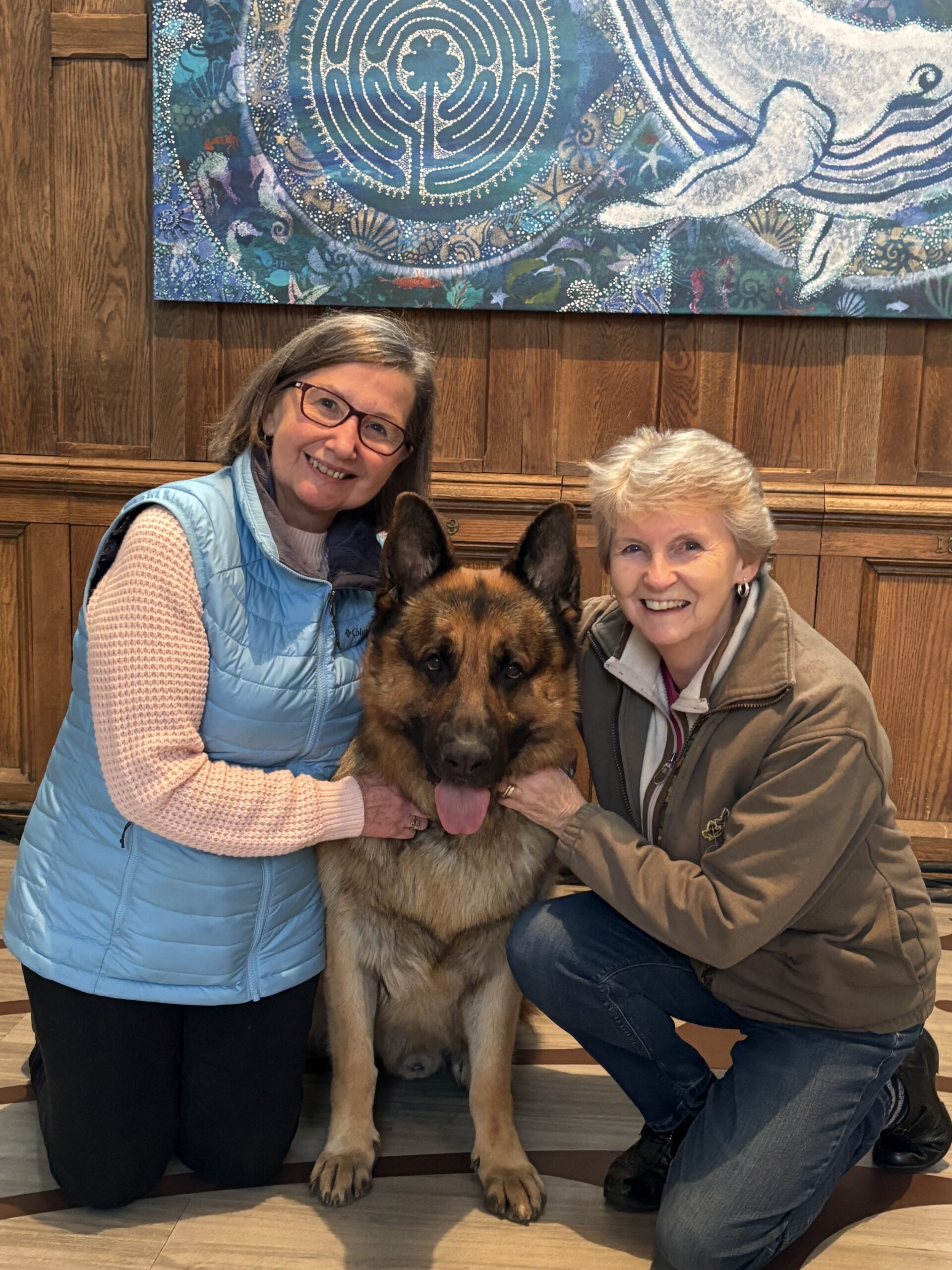 Two women smiling while kneeling with a large German Shepherd in front of wood paneling and a blue nautical-themed mural.