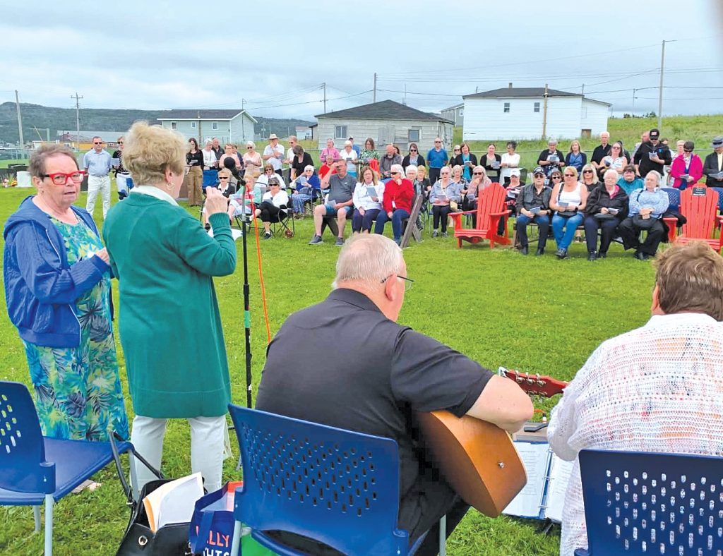 Women speaking into a microphone near musicians at an outdoor gathering.