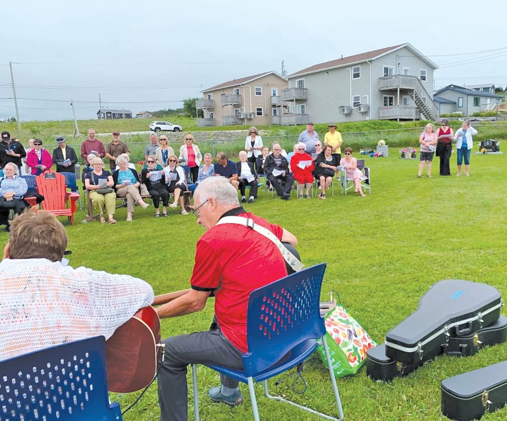 Man playing acoustic guitar for an audience gathered on a grassy lawn.