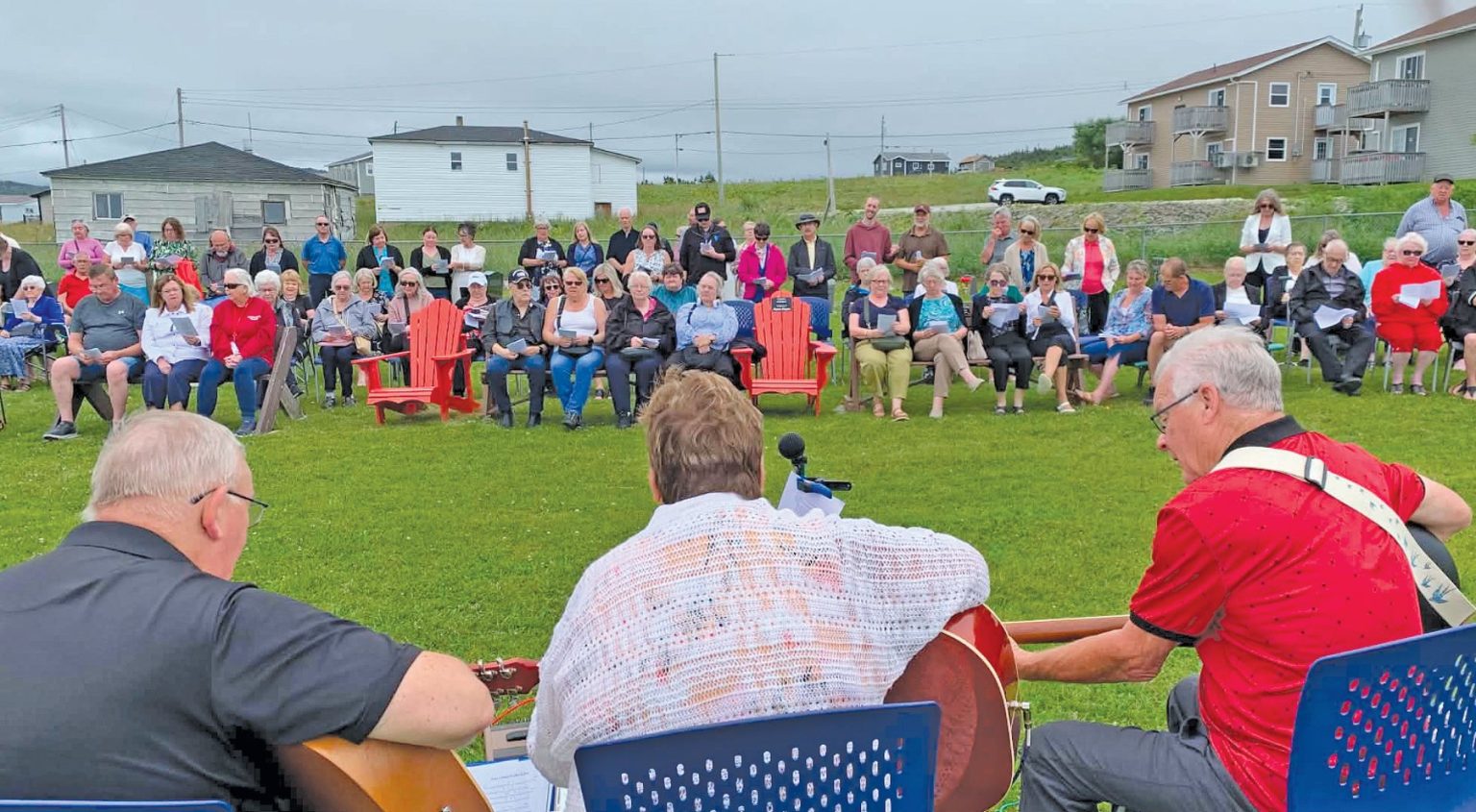 Musicians playing guitar facing a crowd seated outdoors on a lawn.