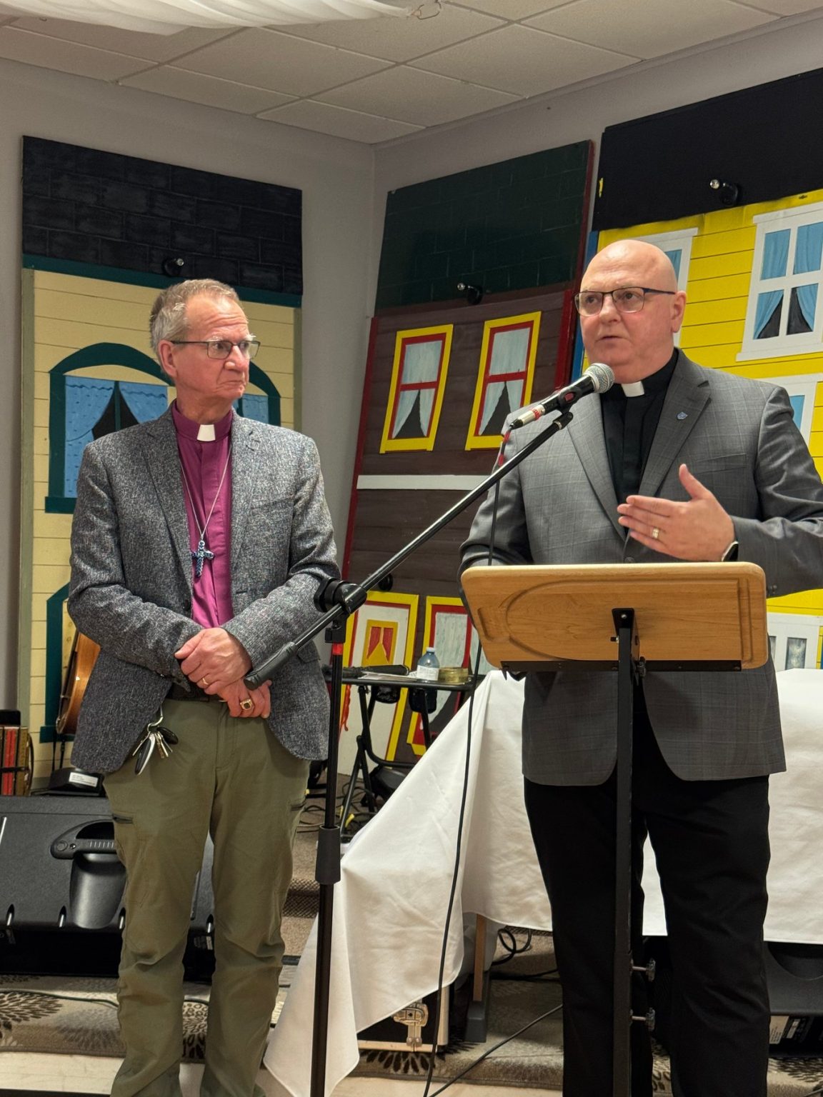 two men, one dressed as a bishop and one as a priest, stand at the front of a room. One speaks into a microphone while the other listens.