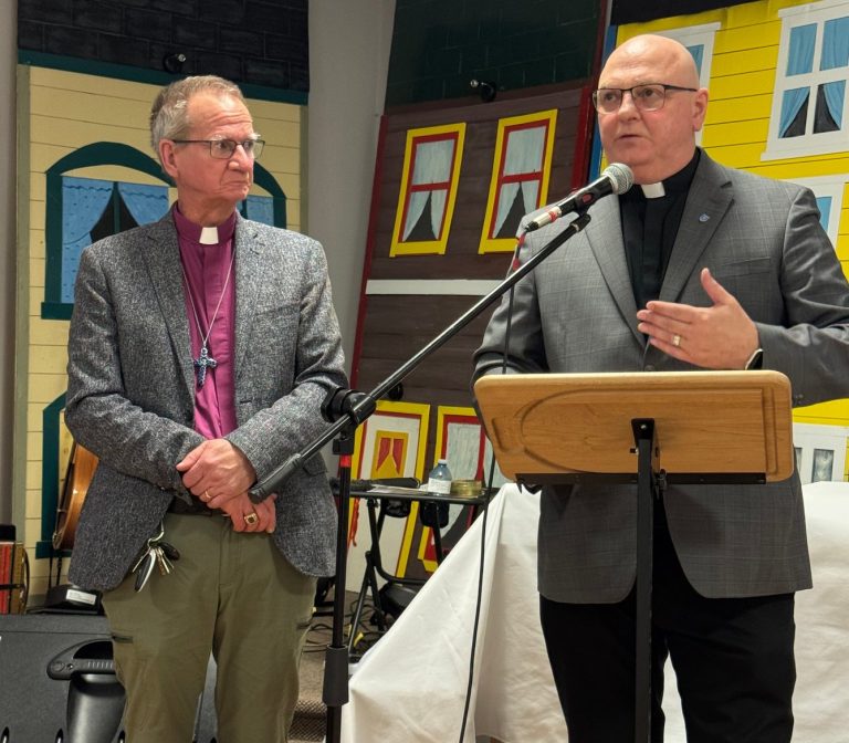 two men, one dressed as a bishop and one as a priest, stand at the front of a room. One speaks into a microphone while the other listens.
