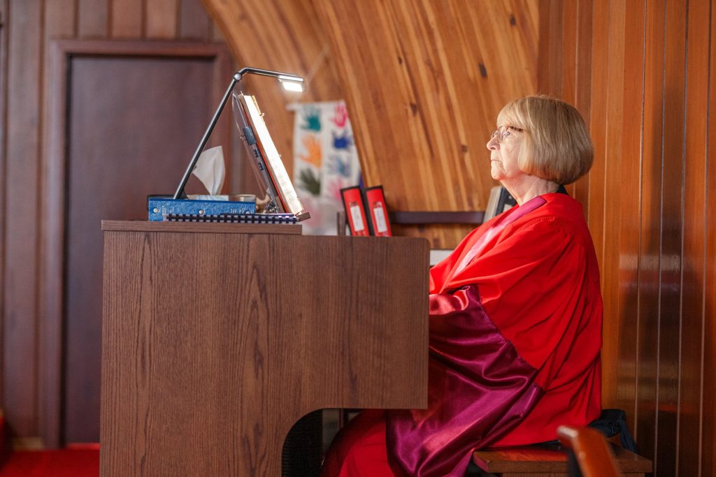 Woman in red academic robes sitting at an organ inside a wooden church.