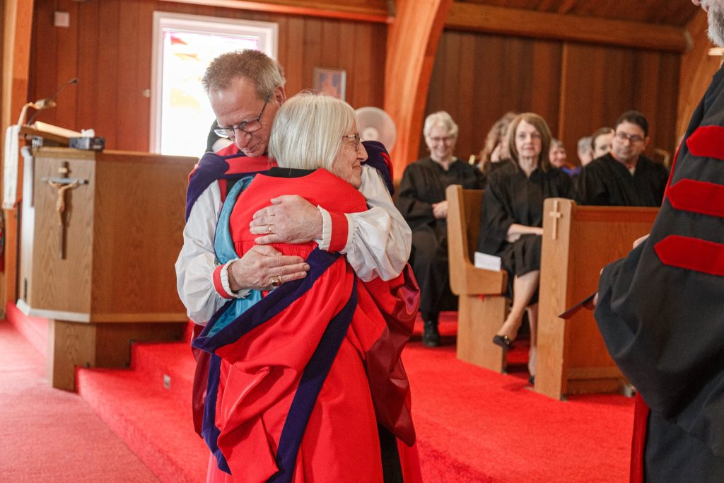 Woman and man in clergy vestments and academic robes smiling together near the church altar.