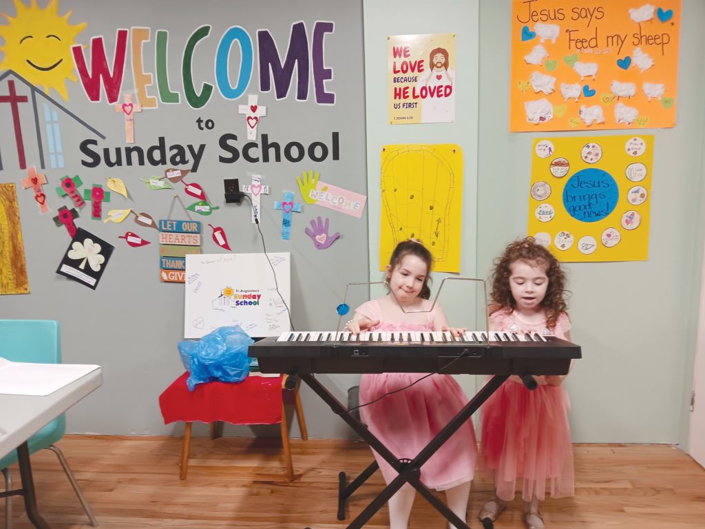 Two young girls in pink dresses playing an electronic keyboard in front of a Sunday School wall.