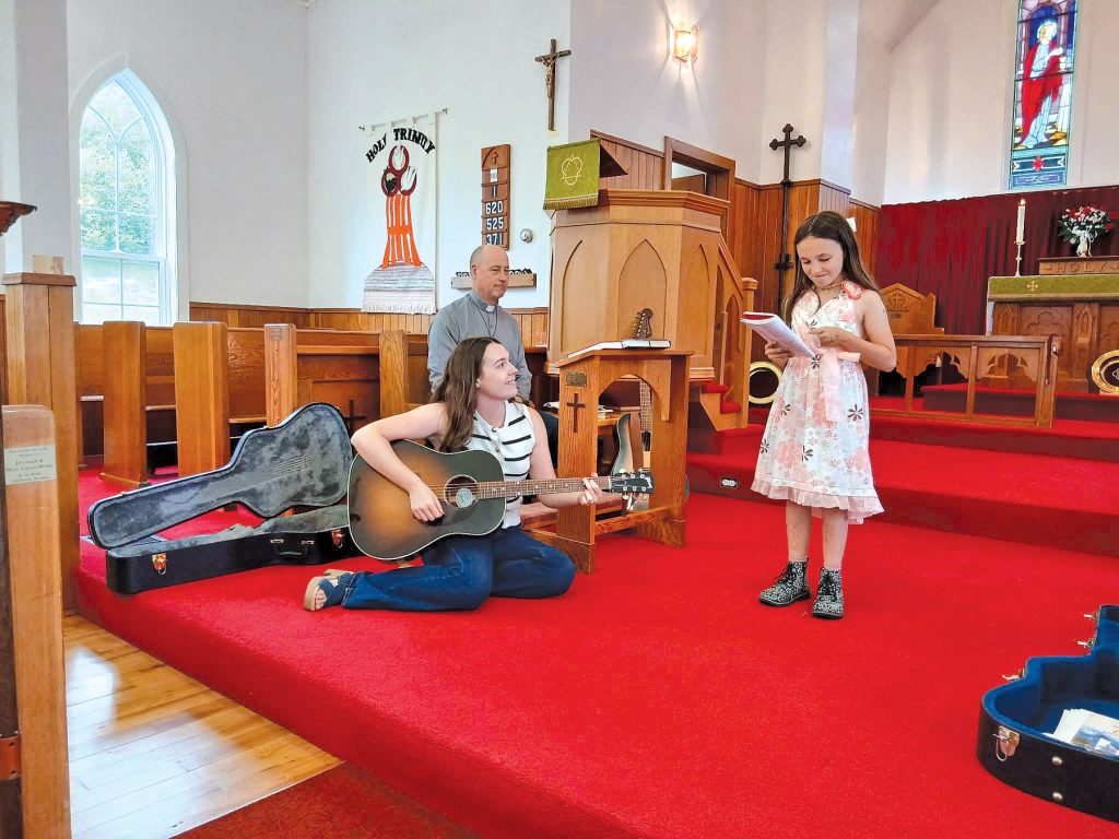 a woman plays the guitar and a girl sings. They are at the front of a church that has bright red carpet.