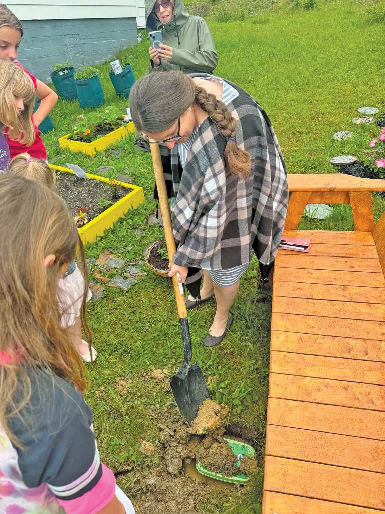 A woman with a log braid down her back digs with a chovel while standing next to a wooden bench. Children watch her.