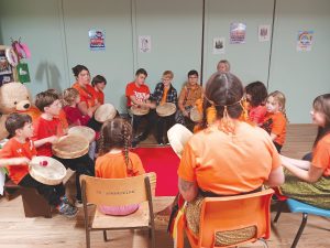 Children sitting in a drum circle playing handheld hand drums together, wearing orange.