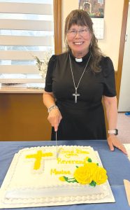 a woman priest cutting a cake that says, "God Bless You Rev Maxine" on it in yellow icing