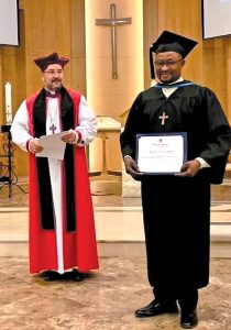 Two men, dressed in clerical and academic gowns, smile at the camera. The man in the front, dressed in black, holds a certificate.
