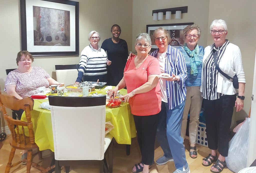 7 women standing and sitting around a table that has a yellow tablecloth on it